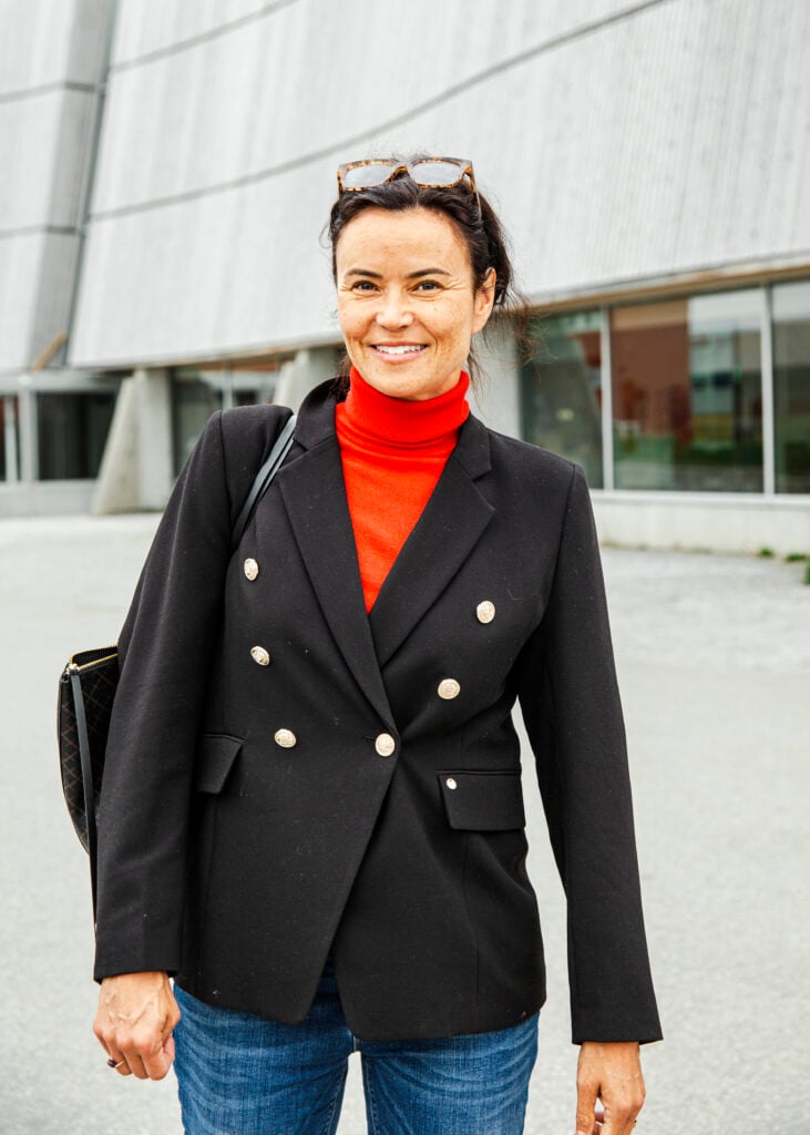 En kvinna i svart kostym och röd tröja ler mot kameran, betong bakom.