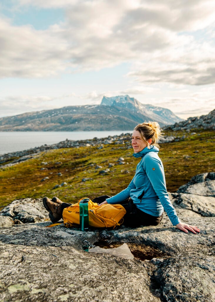 En kvinna i blå tröja sitter ner med en gul ryggsäck och blickar ut över hav och berg.