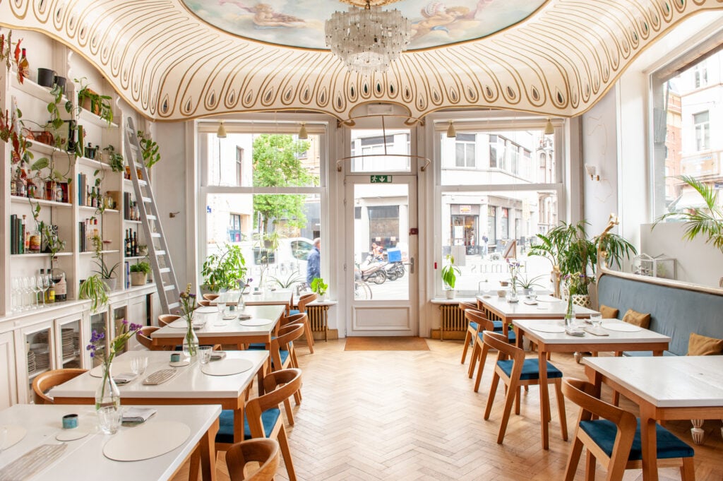 En ljus restaurang med fiskbensparkett, bord och stolar med blåa sitsar. Klassiskt utsirat tag, fönster mot ett torg. 