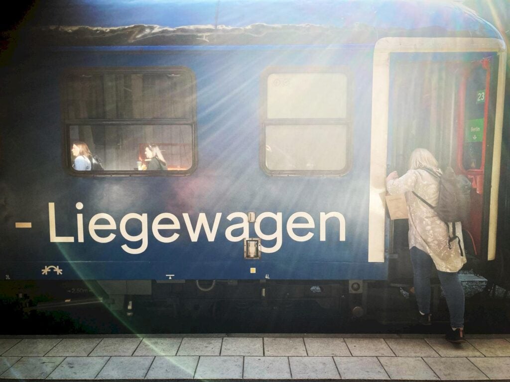 Ett blått tåg med tysk vit text "Liegewagen" på sidan, en person kliver på. Motljus.