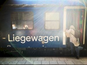 Ett blått tåg med tysk vit text "Liegewagen" på sidan, en person kliver på. Motljus.