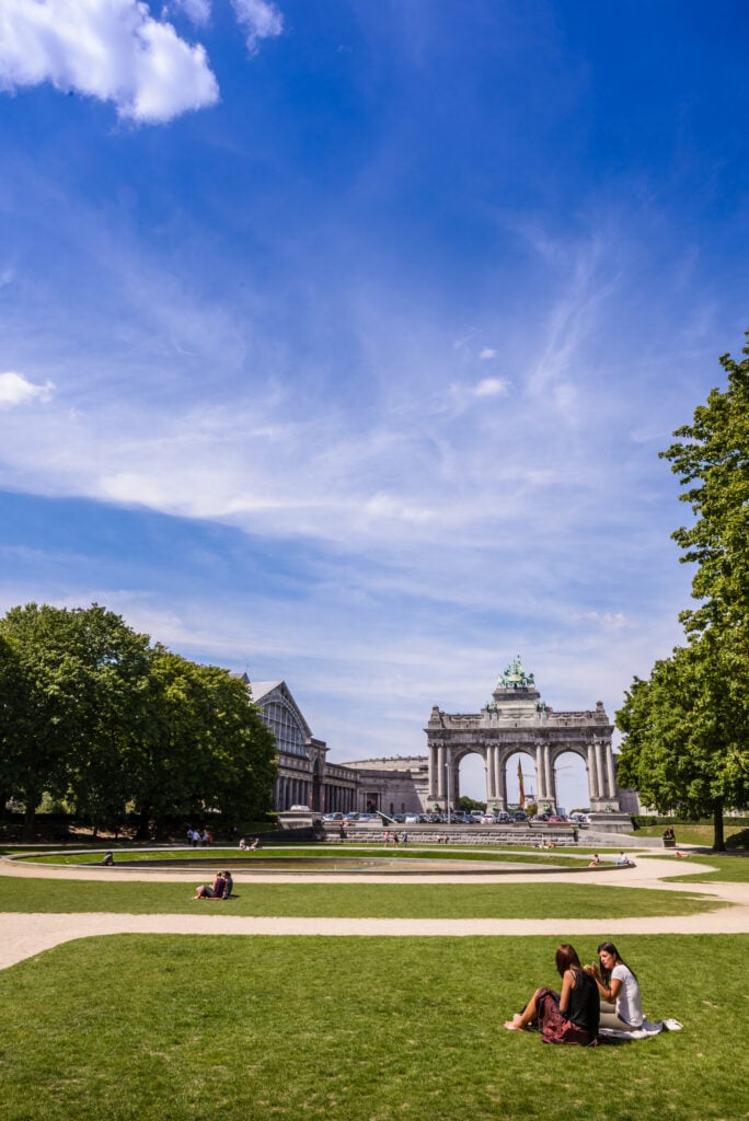 En grön och lummig park med gräsmatta och en triumfbåge. Människor som solar. 