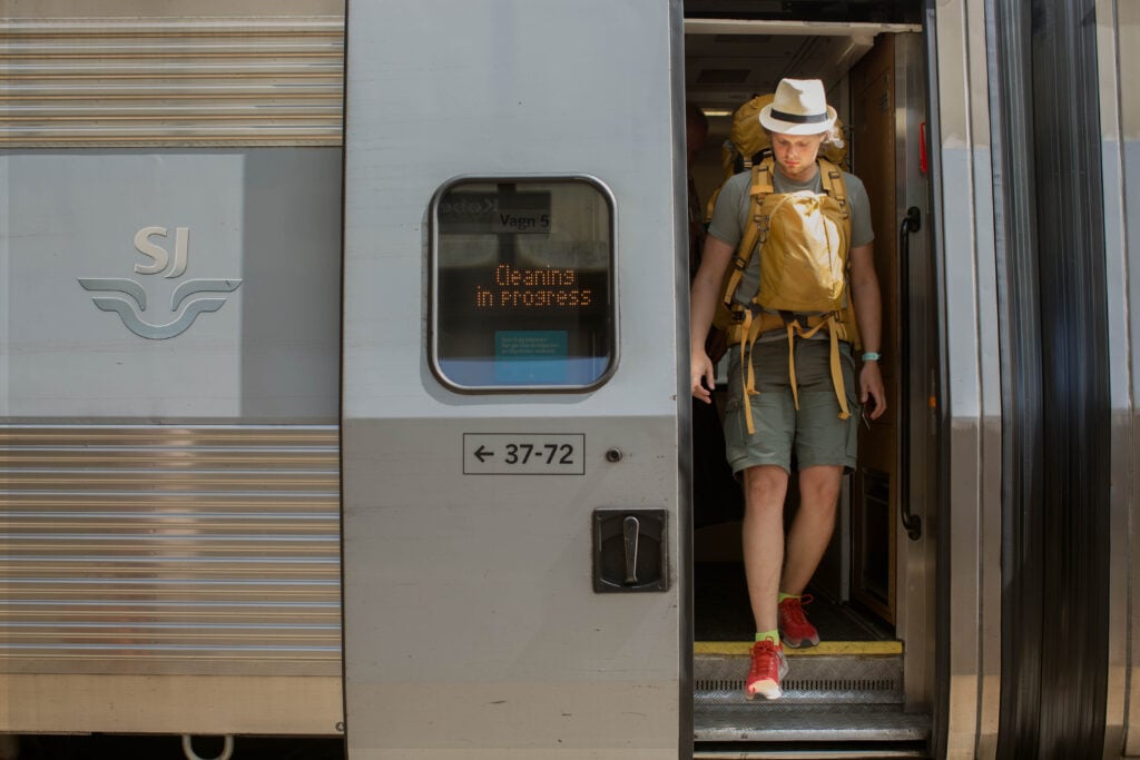 En man i hatt och shorts kliver av ett SJ-tåg med en gul ryggsäck på magen oc på bröstet. 