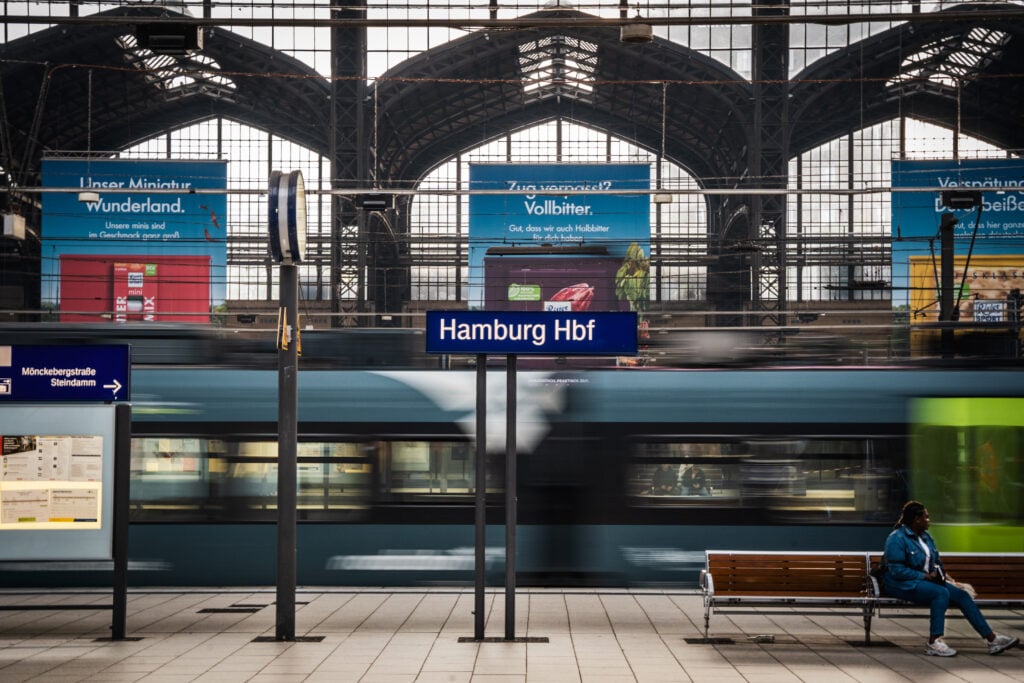 En bly skylt med texten "Hamburg Hbf" syns på en tågperrong, ett tåg susar förbi bakom.