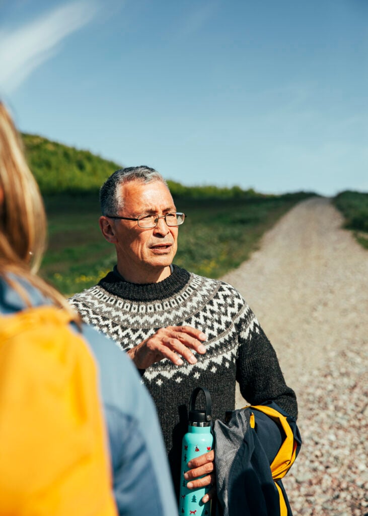 En man i glasögon och stickad tröja kisar i solen på en grusväg.
