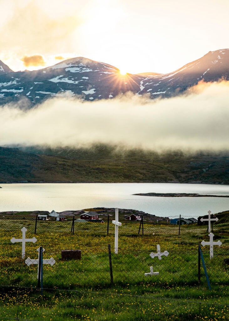 Vita gravkryss på ett fält, framför små hus och ett hav med fjäll bakom.