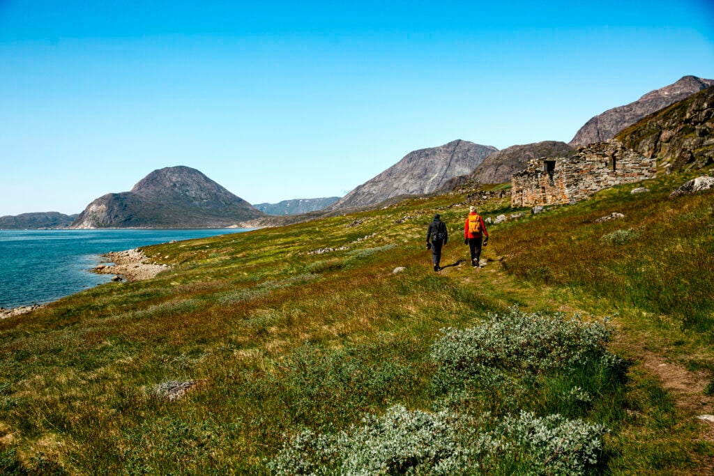 Två personer vandrar över en grön hed, bredvid hav och berg.