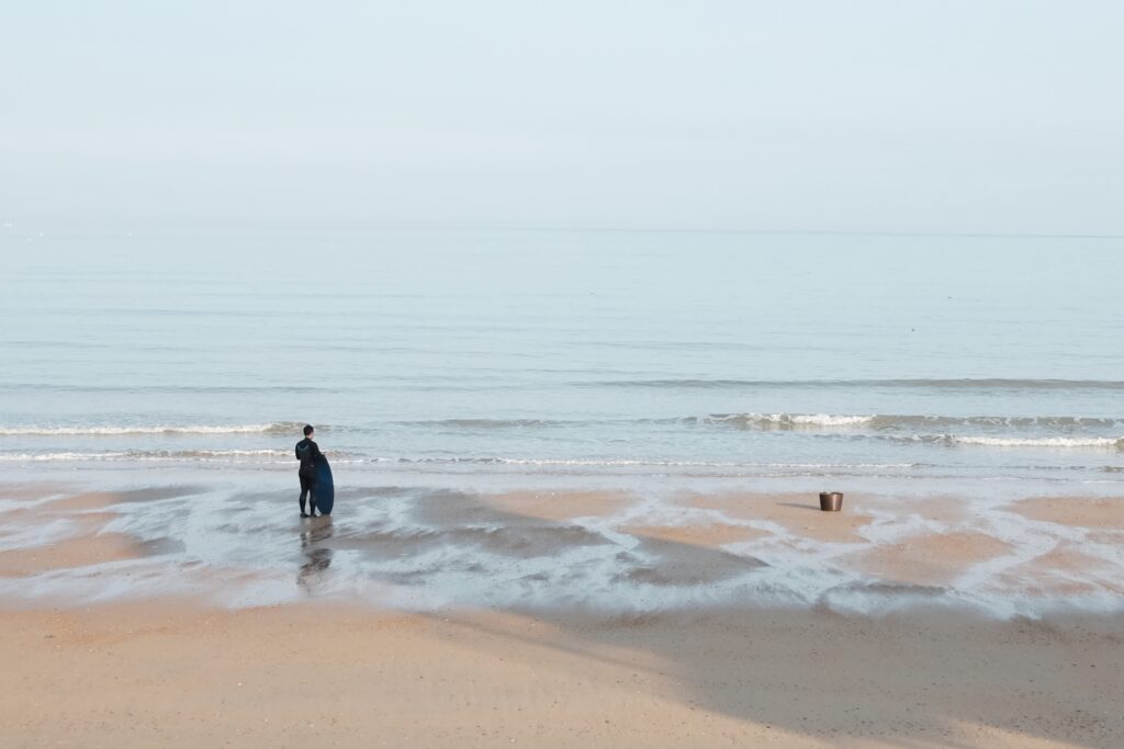 En person står i vattnet på stranden med en surfbräda i handen och blickar ut mot horisonten i havet.
