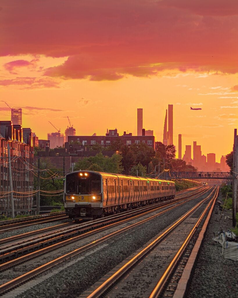 Ett tåg i solnedgången på en järnväg. I bakgrunden syns en stad och ett flygplan. 