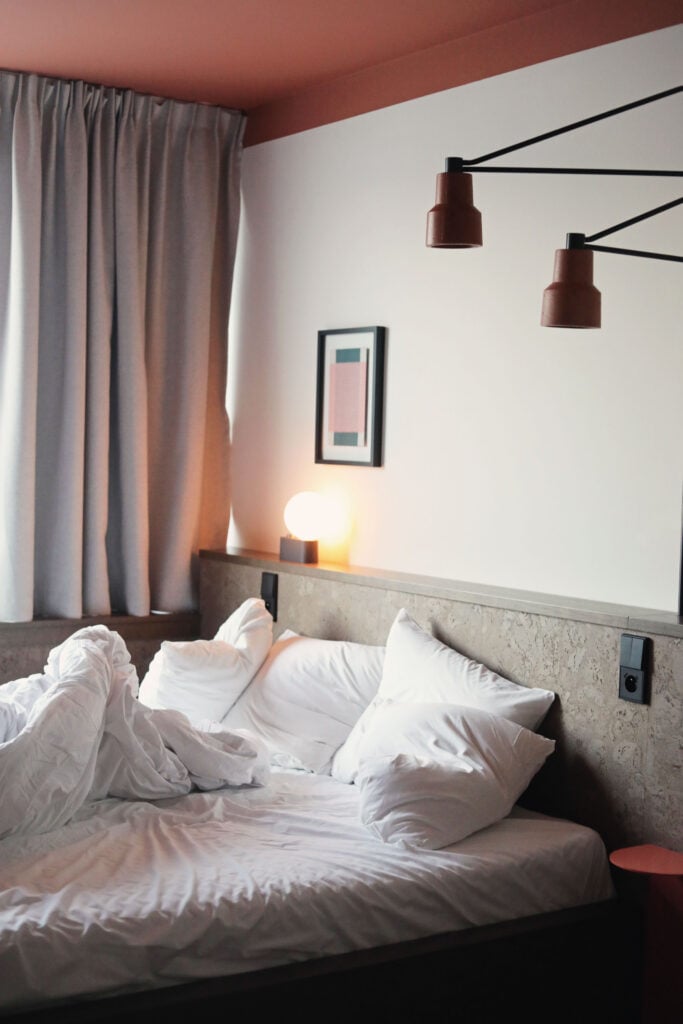 En grå hotellsäng med vita täcken och kuddar, vit vägg, grå gardiner. 