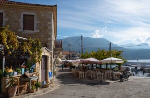 En liten servering på kajkanten med små stenhus bredvid och båtar som guppar. Dramatiska berg i bakgrunden,