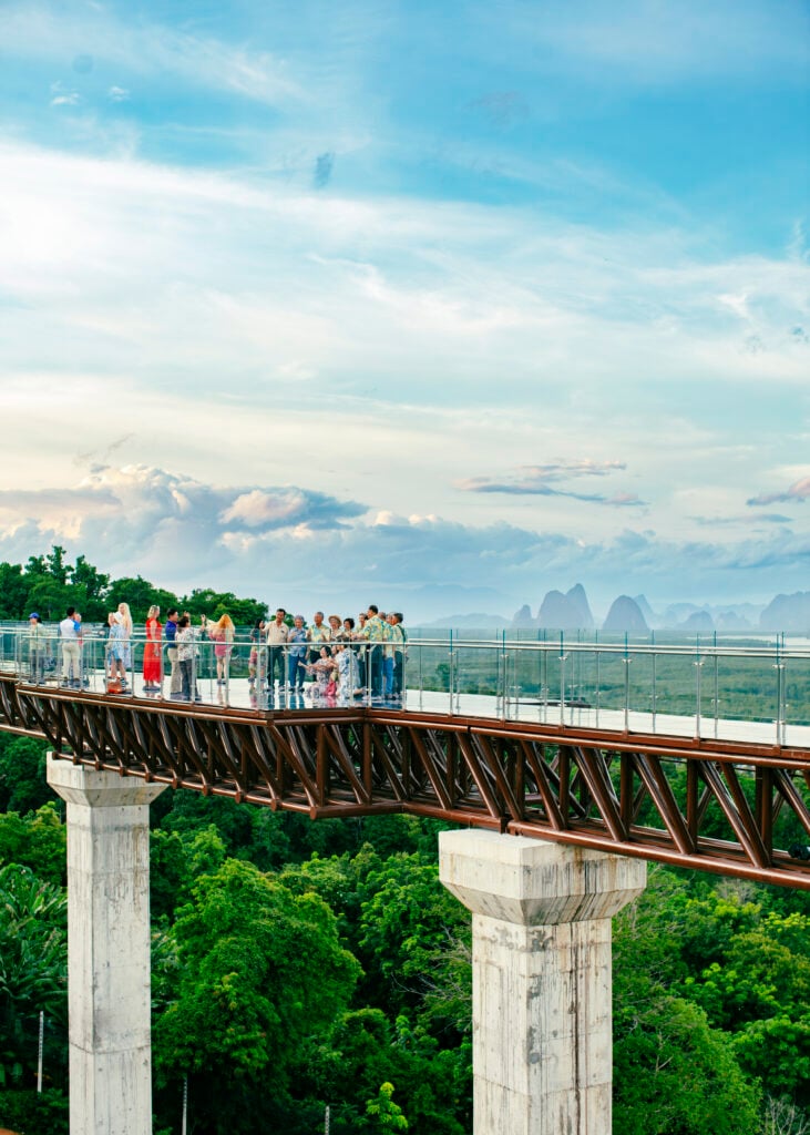 En turistgrupp står på en glasbro högt ovan djungeln, mycket dramatiska berg bakom.
