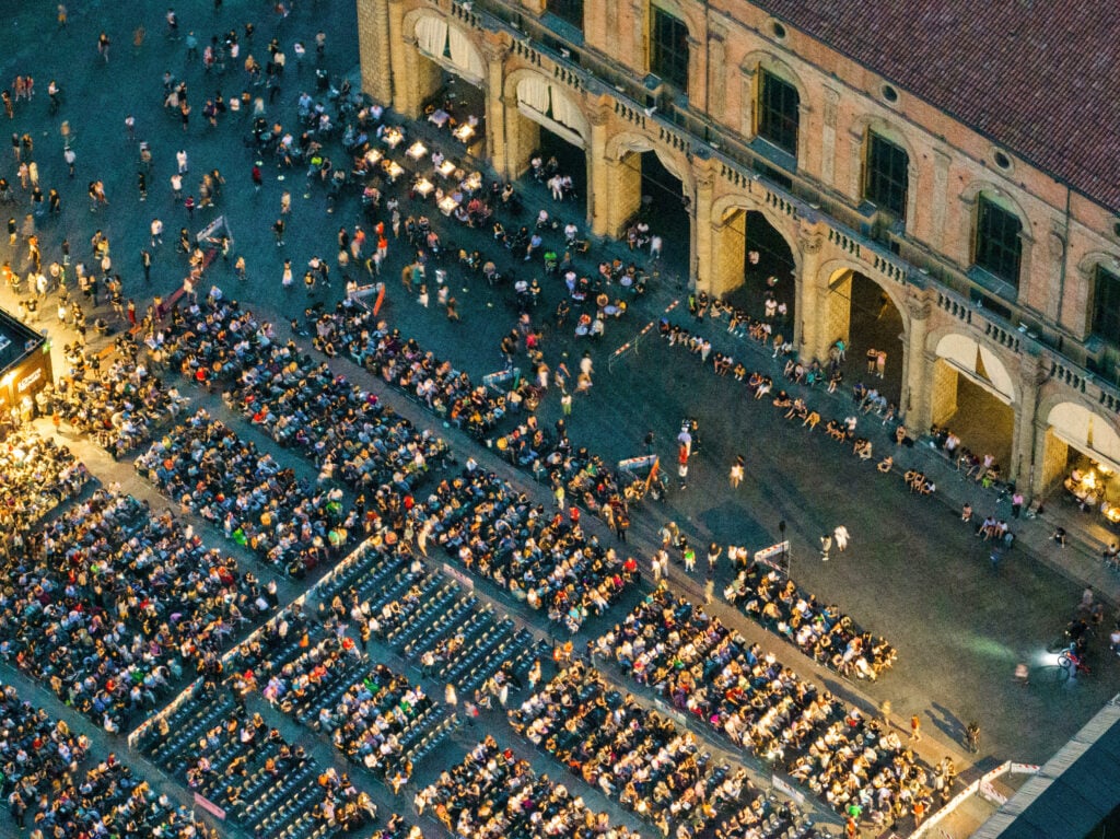 Massor av människor sitter i rader på stolar på ett torg. Drönarvy.
