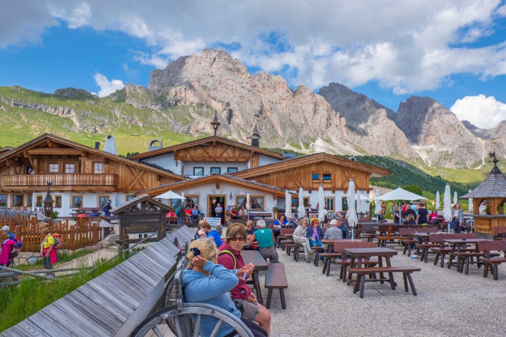 Turister sitter på en skuggad uteservering under bergen, hus i vitt och korsvirke.