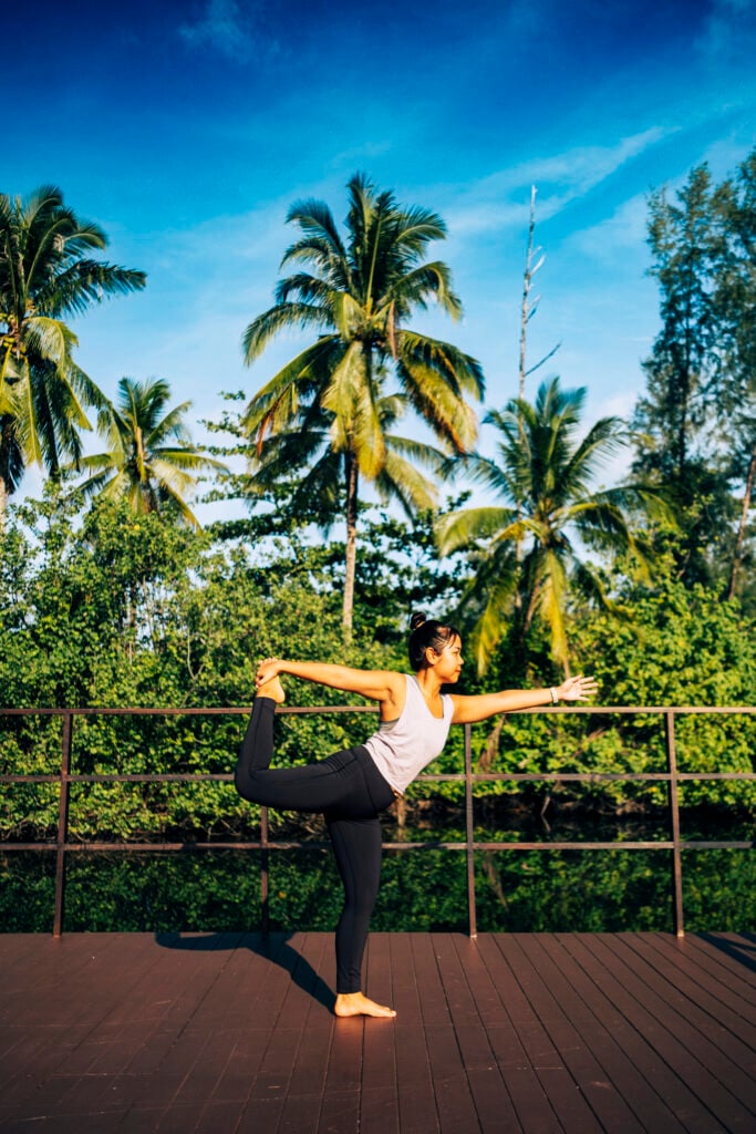En thailändsk kvinna med vitt linne och svarta tajts står och balanserar på ett ben i en yogapose, framför en grön djungel.