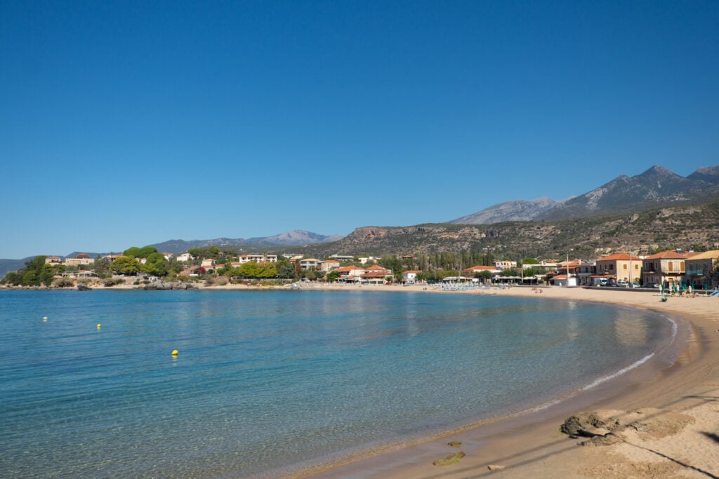 En tom strand vid turkost hav, hus och berg i bakgrunden. Blå himmel. 