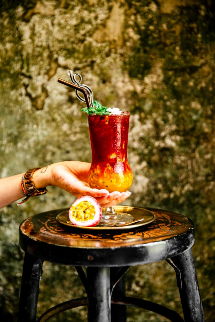 En röd och gul fruktdrink i ett glas med sugrör, en hand håller i glaset.