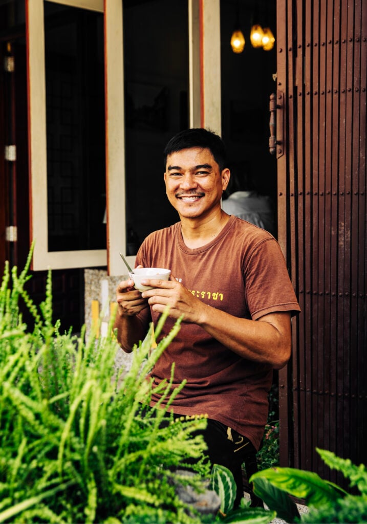 En thailändare i brun tröja håller i en kaffekopp utomhus.