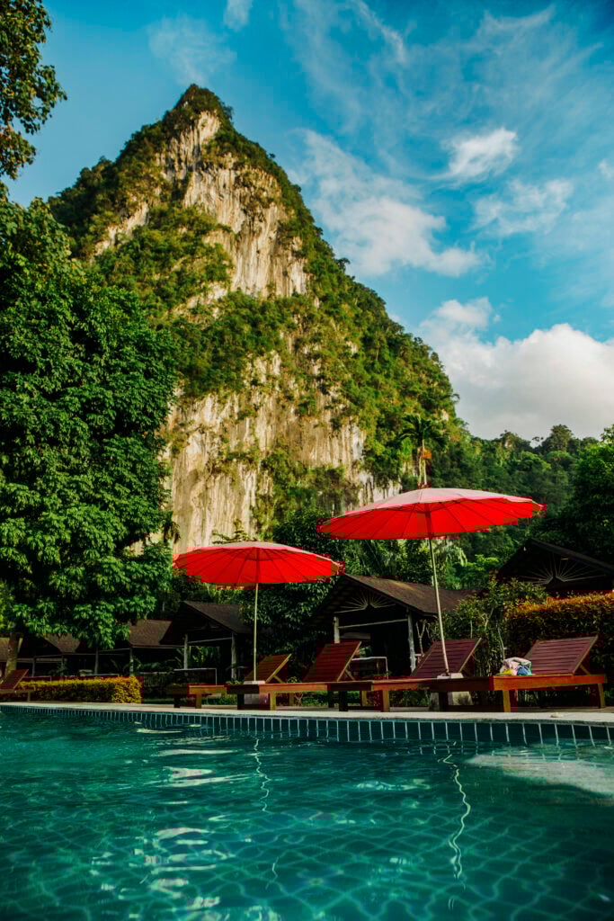Turkost poolvatten bredvid solsängar med röda parasoll under grönskande berg.