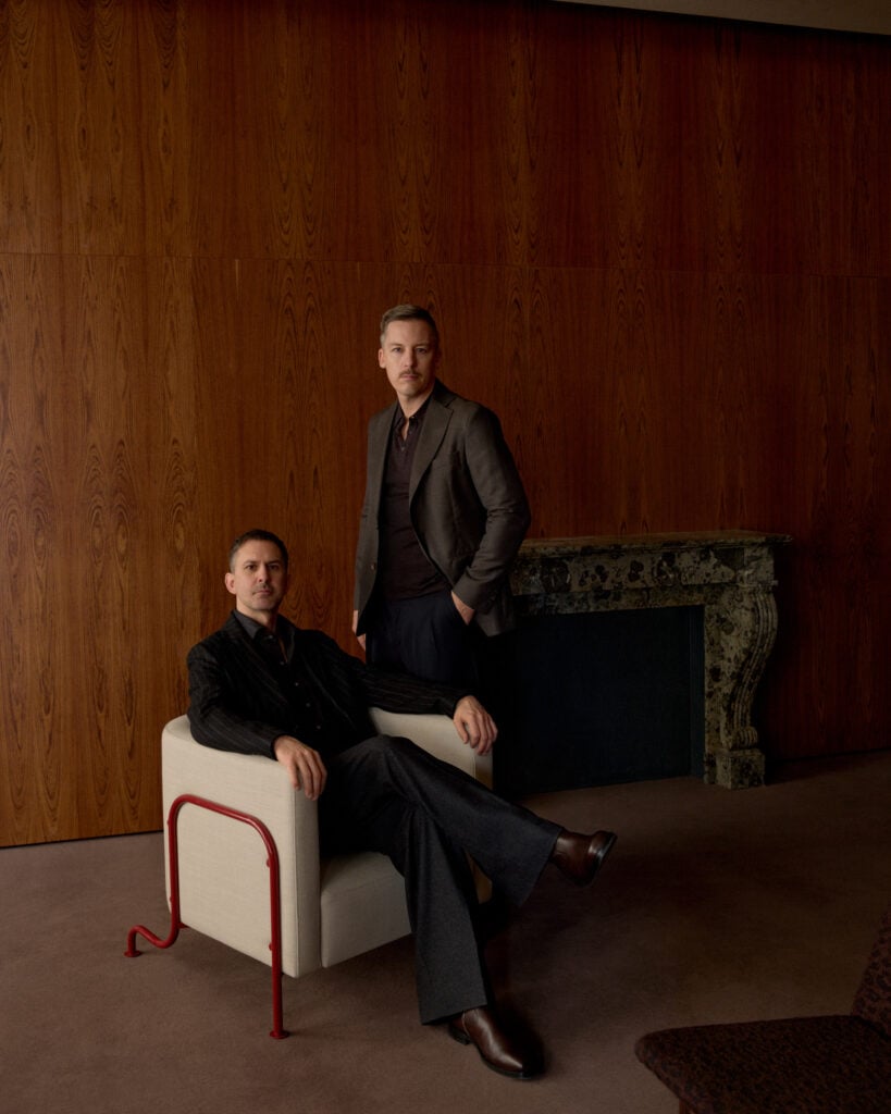 Två män poserar framför kameran, den ena sitter i en vit fåtölj, den andra står bredvid. 