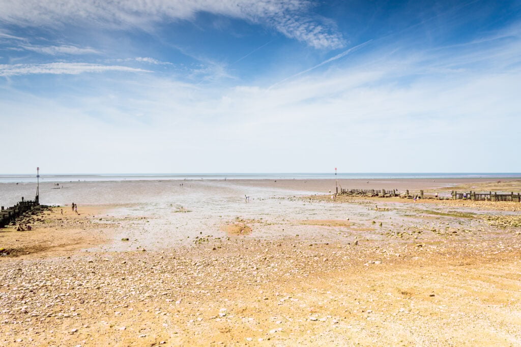 En beige strand sticker ut i havet, små pålar syns i horisonten. Blå himmel. 