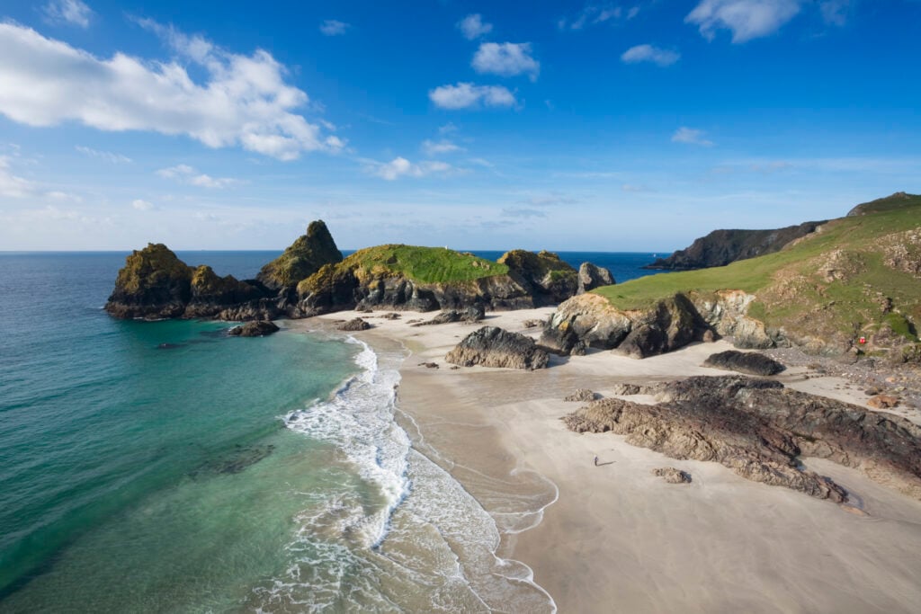 En vit sandstrand möter turkost vatten, vackra kalkstensklippor tornar upp sig. Gröna hedar syns på sidan. Klarblå himmel. 