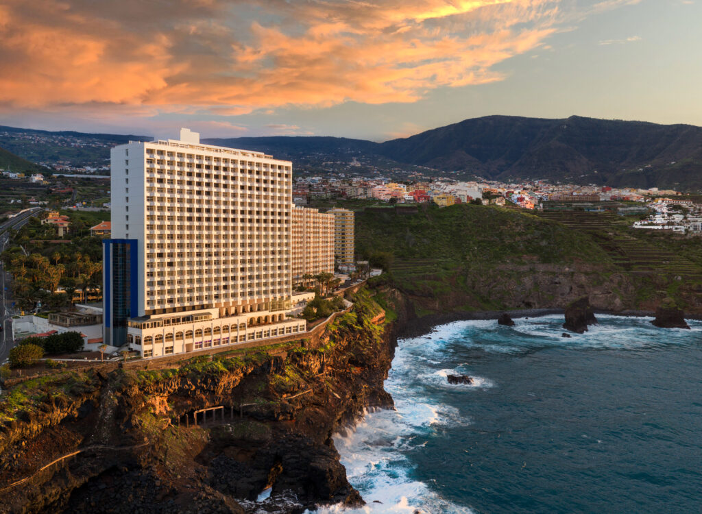 Ett stort högt hotellkomplex reser siv ovanför vulkaniskt svarta klippor och blått hav, på himlen syns gyllene moln. 
