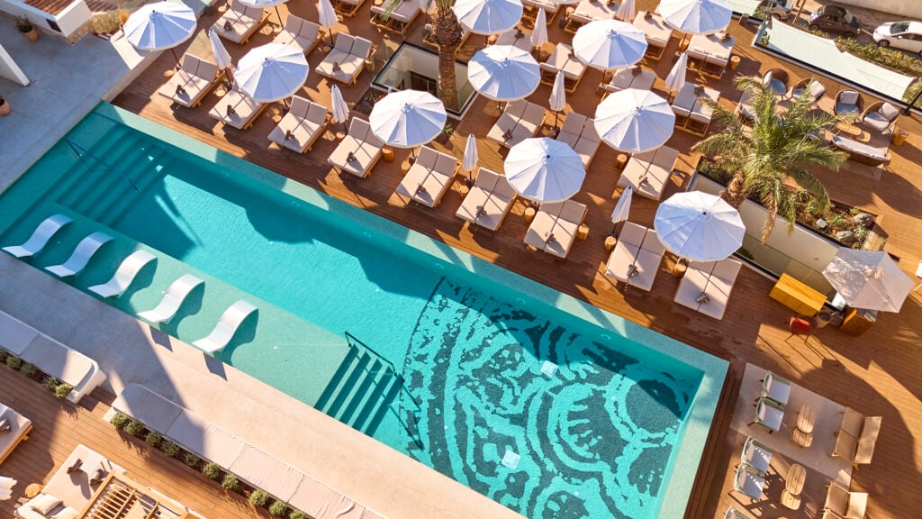 Drönarvy ovanför en avlång fyrkantig pool med turkost vatten och ett svart mönster på botten. Ett teracottafärgat pooldäck, med massor av solstolar och vita parasoller. 