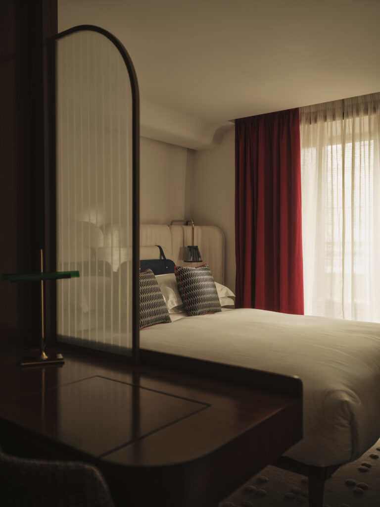 En säng med vita lakan, randiga kuddar, framför ett fönster med röda gardiner, mjukt ljus skiner in i rummet. 