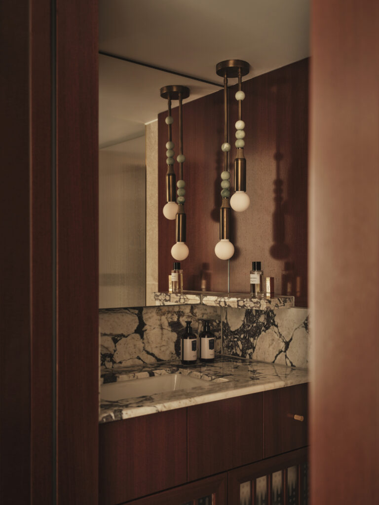 Ett litet badrum syns byggt i mörkt trä. en svart/vit marmorskiva, designade lampor hänger ner framför spegeln. 
