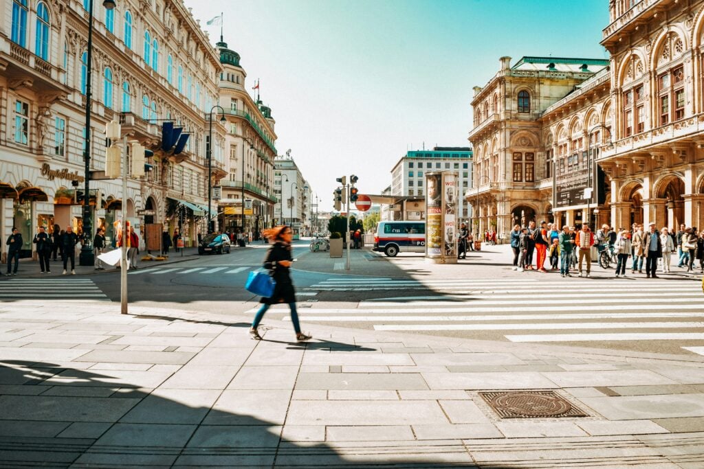 Människor går i solen mellan klassiska stadsbyggnader i Wien, blå himmel. 