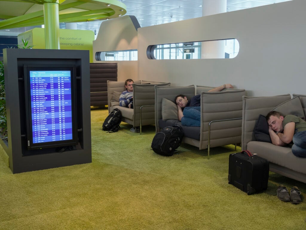 Personer sitter och sover i väldigt ombonade grå sovfotöljer. Bakom finns syns bokningsbara sovkuber. En snorgrön heltäckningsmatta fyller rummet. 