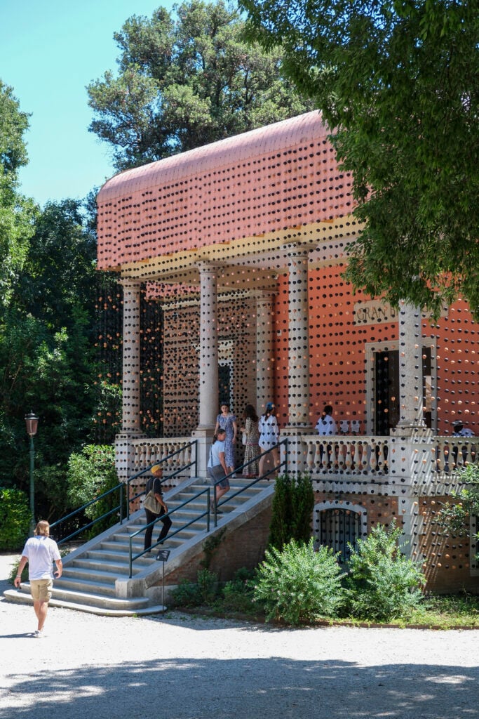 En klassisk, rosa fasad med kolonner, en trappa upp, många turister går in. Små svarta och rosa bollar hänger i ett draperi runt hela huset. 