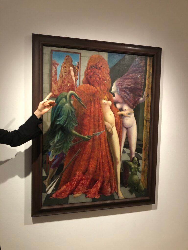 En hand pekar på en målad tavla med nakna människor och en grön fågel. 