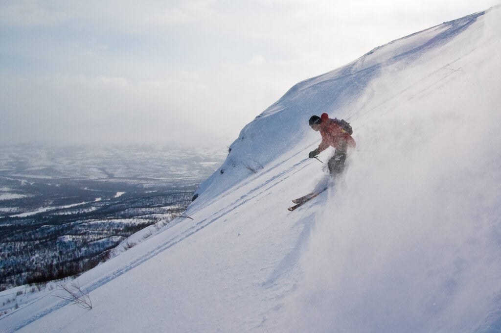 En kille i röd jacka åker skidor nedför ett pudertäckt berg. 