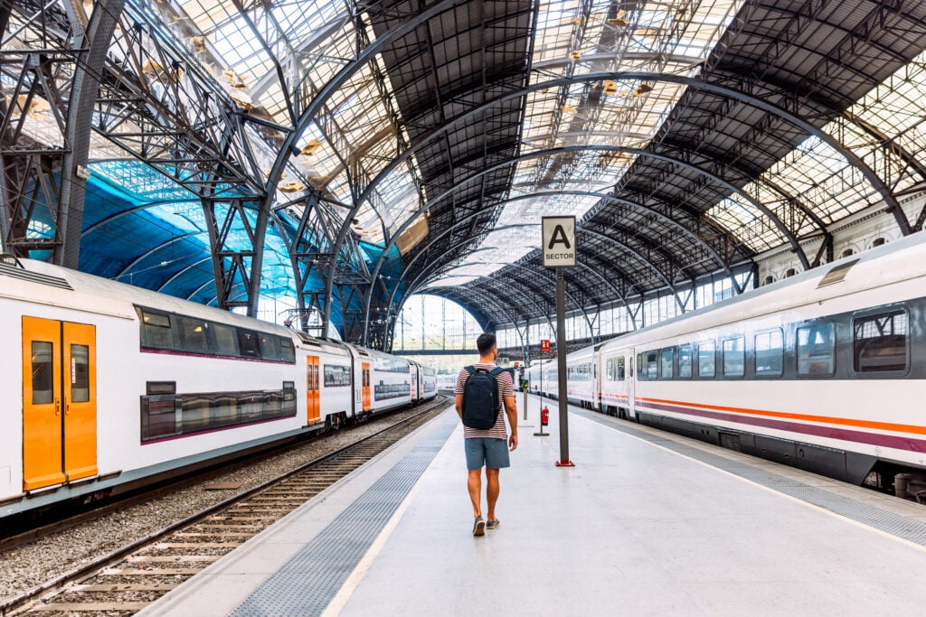 En man med ryggsäck går längs med en tågperrong, ljuset flödar in genom glastaket. 