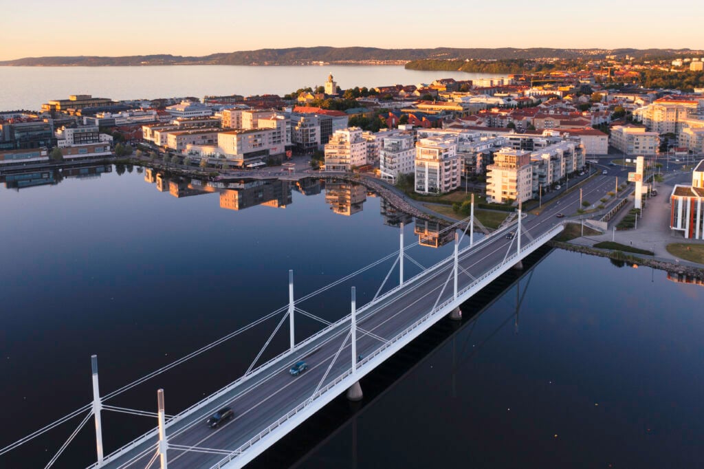 Drönarbild över spegelblanka vatten och en stad i solnedgången, en lång bro går rakt över en sjö.