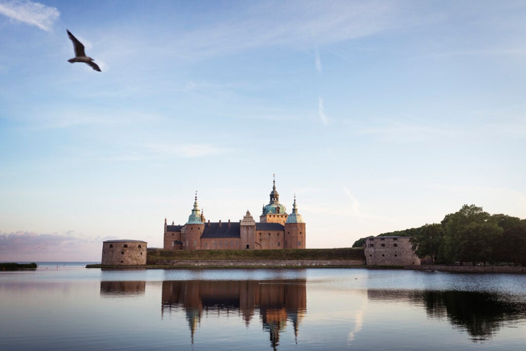 Ett slott i solnedgången bredvid spegelblankt hav och ljus pastellblå himmel. 