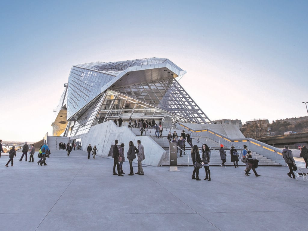 En vit, kantig byggnad med mycket glas, på ett grått stilrent betongtorg. Välklädda människor pratar med varandra. 