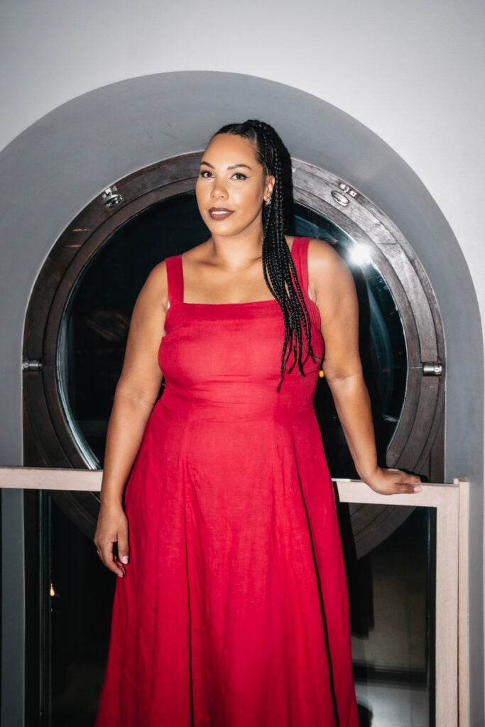 En mörkhyad kvinna med svart flätat hår i röd klänning ler mot kameran.