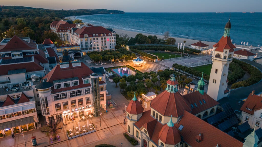 Flygvy ovanför ett torg med klassiska byggnader, havet skymtas bakom.