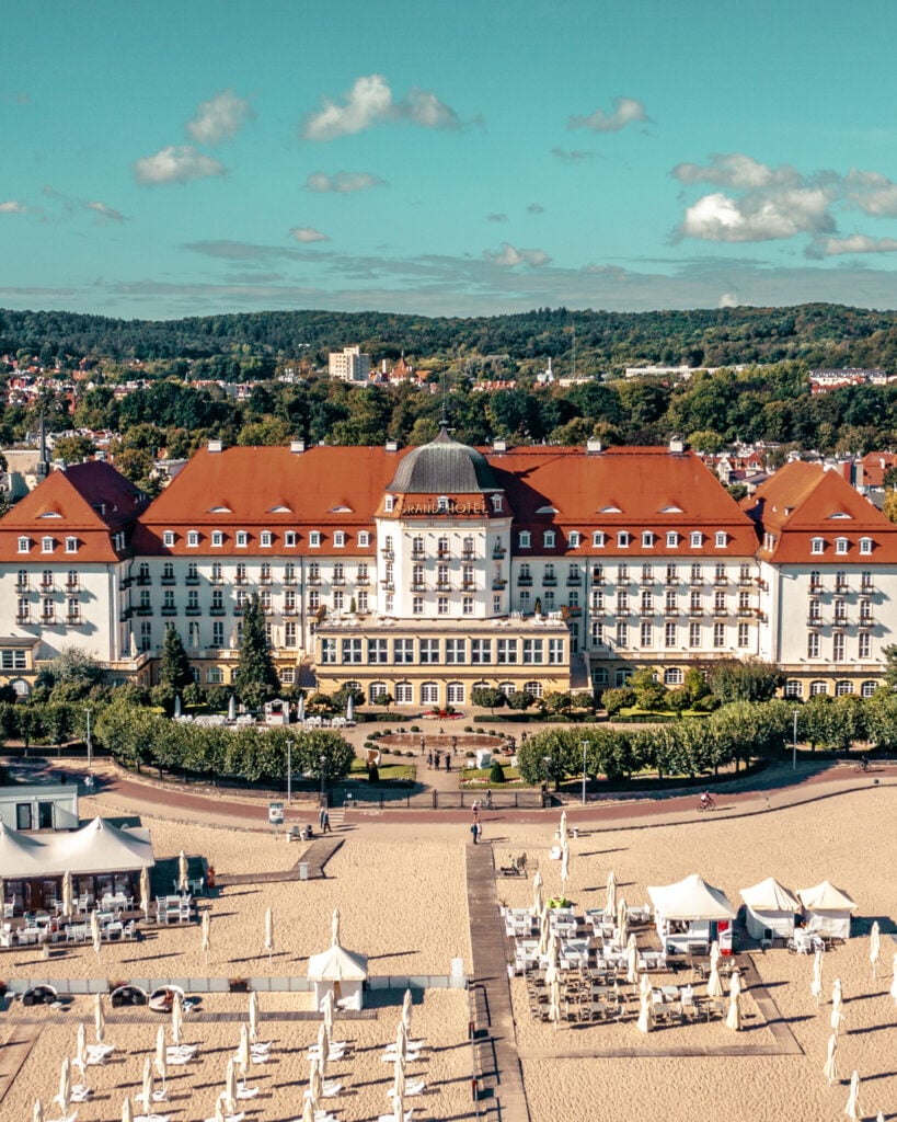 En magnifik hotellbyggnad med vit fasad, rött tak, två flyglar, en halvmåneformad trädplantering framför.