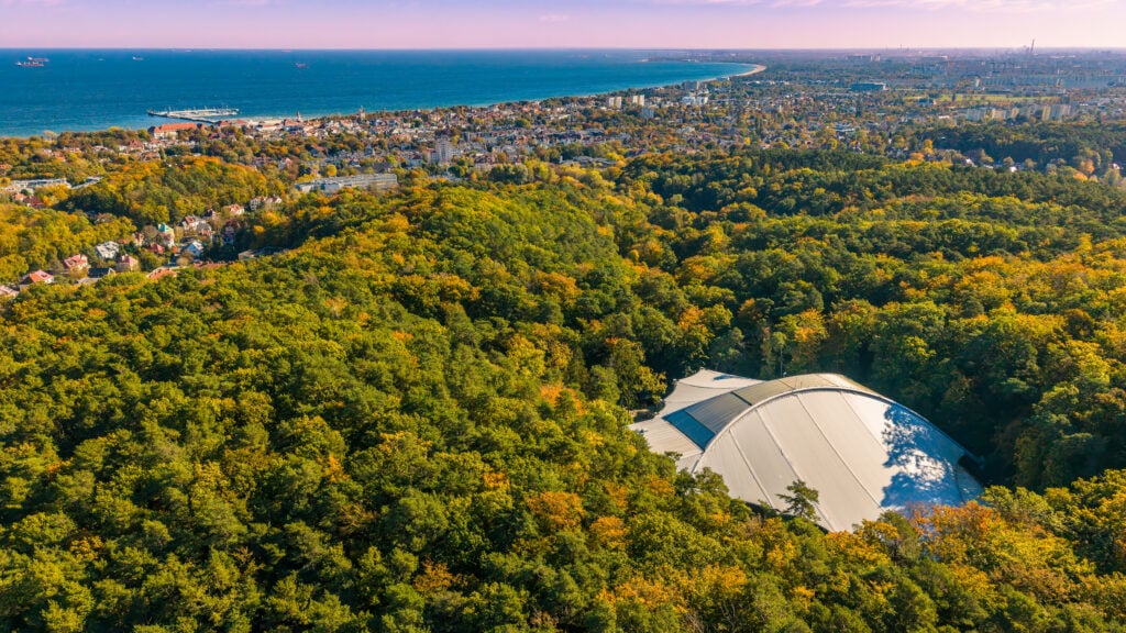 En välvd byggnad bland träden i skogen, havet och en stad i bakgrunden.
