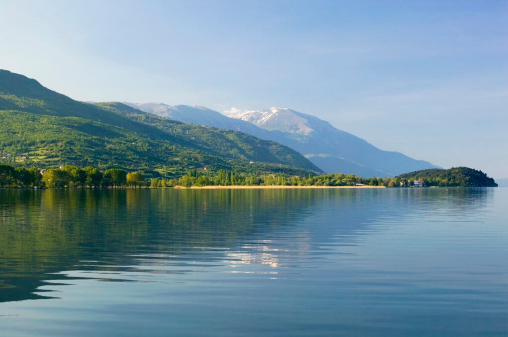 En spegelblank blå sjö framför höga berg, täckta med lummig grönska. 