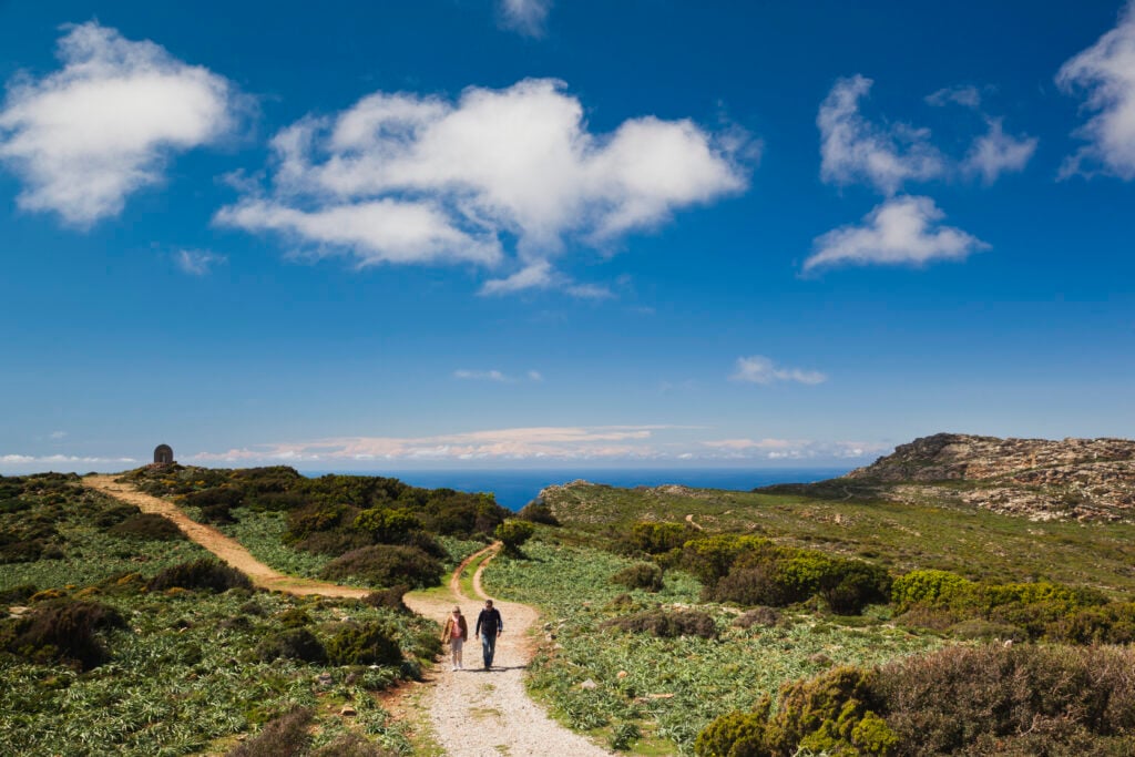 Två personer promenerar på en kalkgrusad stig som slingrar sig fram bland låg grön vegetation, långt borta syns havet. 