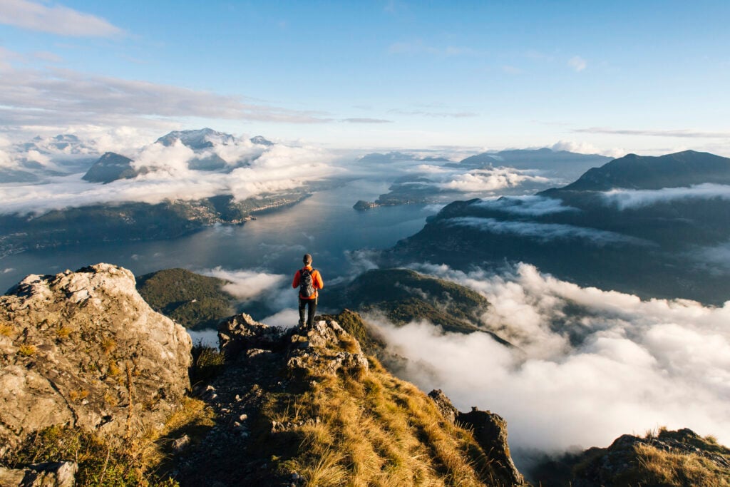 En person i orange jacka och ryggsäck står på en bergstopp och blickar ut över en sjö. Molntussar på himlen. 