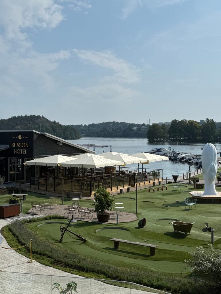 En grön gräsmatta och vita parasoll omgärdar en mörkgrå kantig hotellbyggnad framför vattnet. 