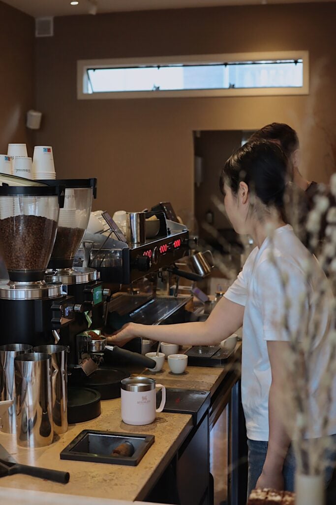 En kvinna med svart hår i knut står bakom en disk och hanterar massor av avancerade kaffemaskiner. 