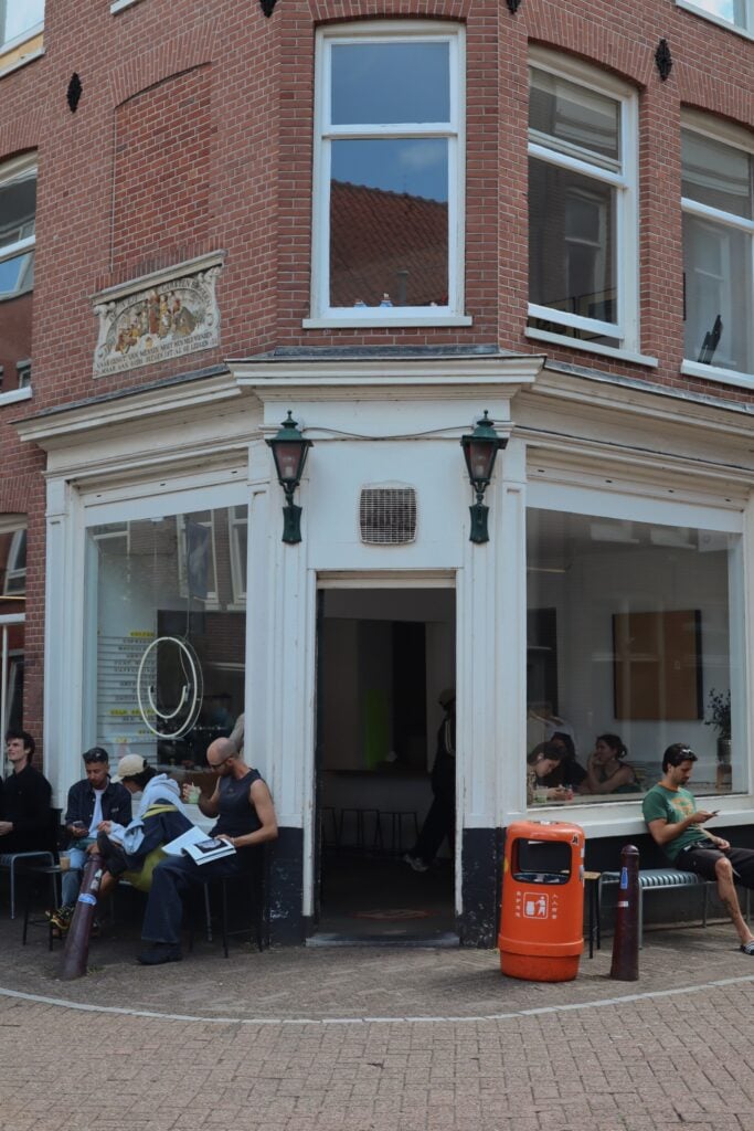 Ett gathörn med ett café med stora fönster, knallröd papperskorg utanför. människor sitter på bänkar och dricker kaffe. 