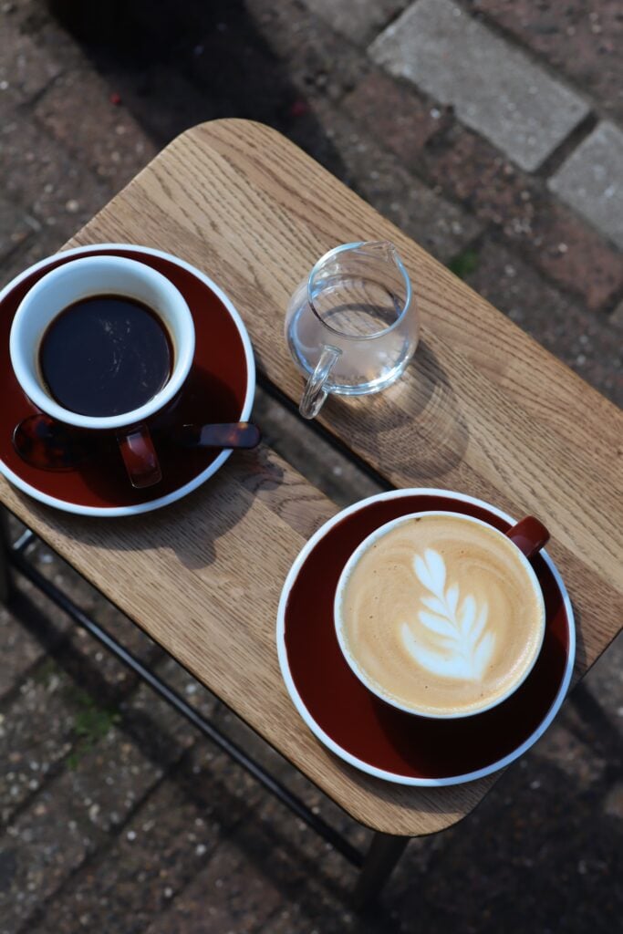 Två röda kaffekoppar med fat, en med svart kaffe, en med latte. Ett glas vatten. Fotat ovanifrån. 